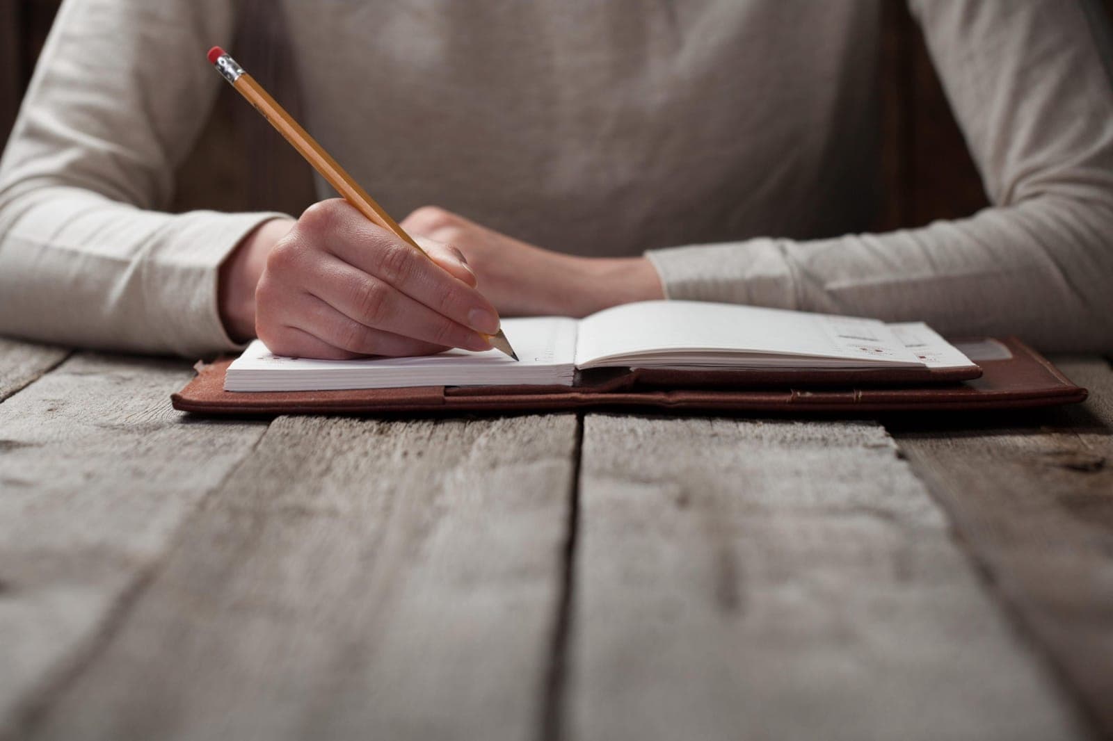 Hands writing with a pencil in an open notebook on a wooden table